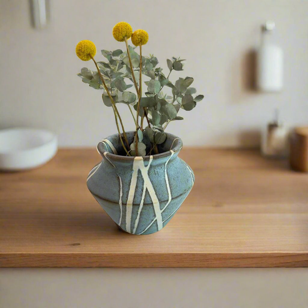 
                      
                        Medium sized vase with white lines in random pattern against a matte light blue background with flowers
                      
                    