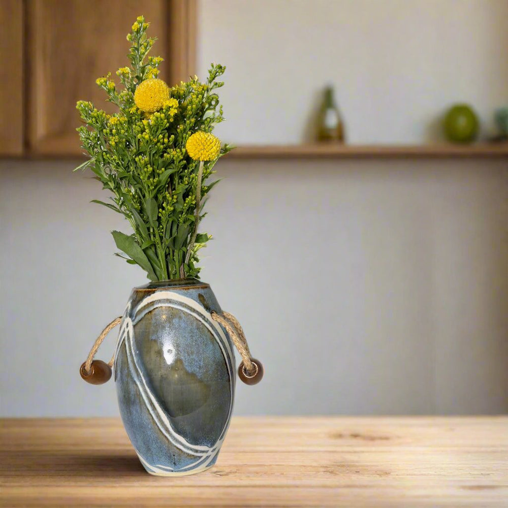 Vase with jute handles and wooden beads filled with yellow flowers. Glazes are brown and blue with white lines