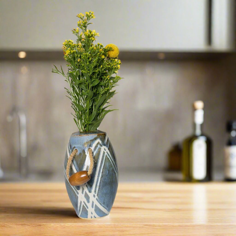 
                      
                        Vase with jute handles and wooden beads filled with yellow flowers. Glazes are brown and blue with white lines
                      
                    