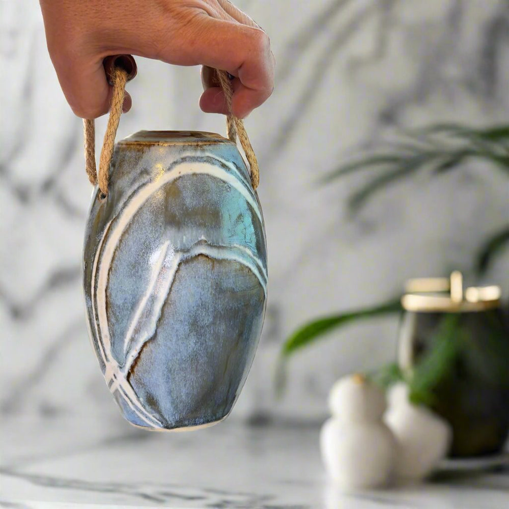 
                      
                        Hand holding Vase with jute handles and wooden beads. Glazes are brown and blue with white lines
                      
                    